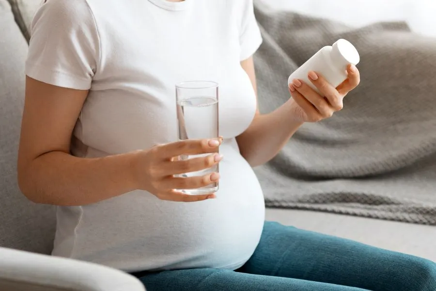 A pregnant women sits on a couch and reads the ingredients on a supplement bottle.